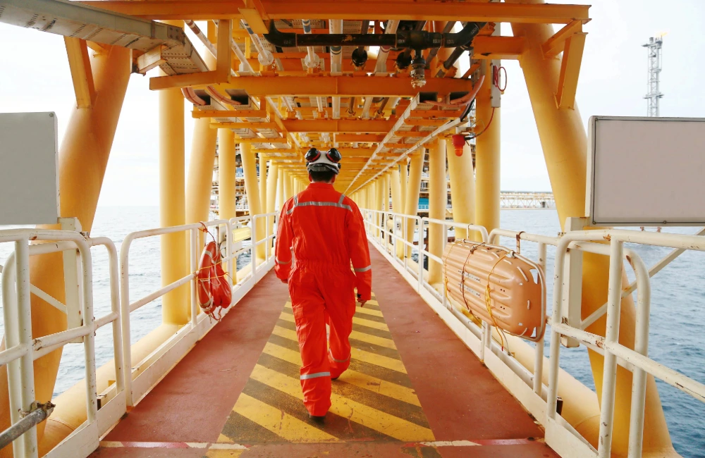 An offshore oil and gas worker in orange overalls walks on an oil rig.