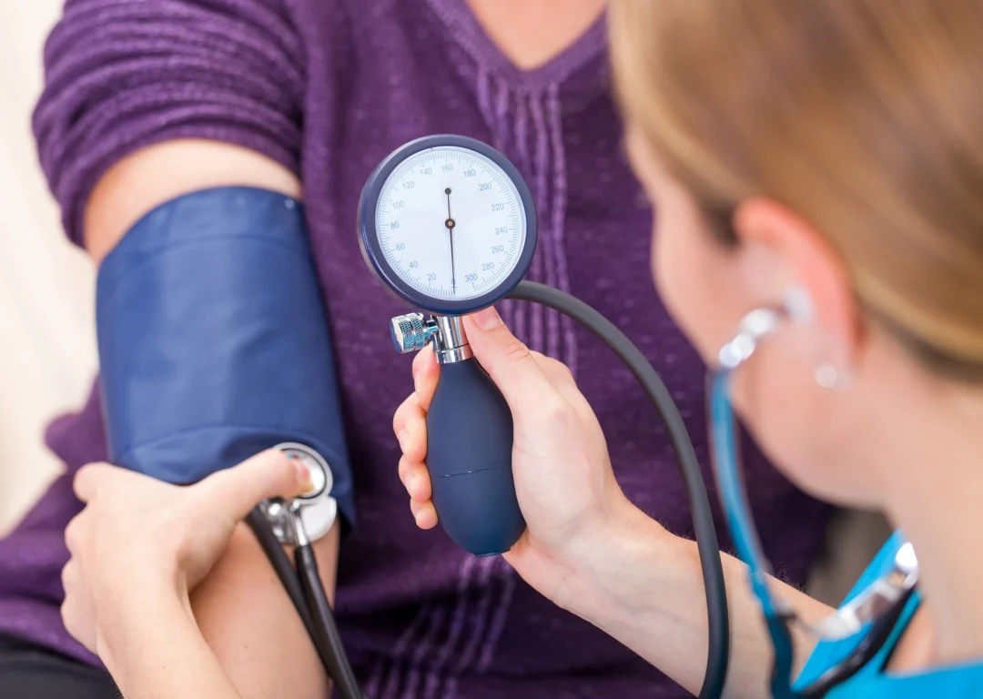 A woman in a purple top having her blood pressure taken by a female doctor.